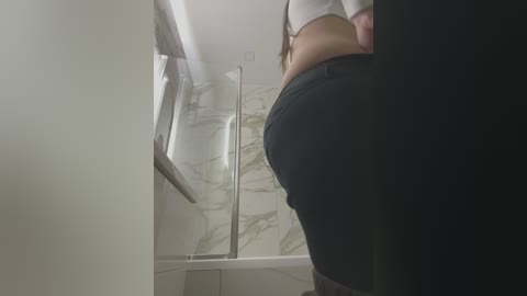 Media: Video of a woman with a curvy figure, wearing tight black jeans and a white top, standing in a modern bathroom with marble tiles and a glass shower enclosure.