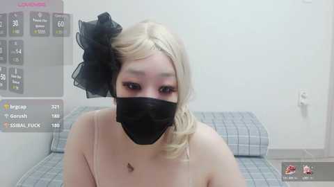 Media: A video of a young Asian woman with fair skin and platinum blonde hair, styled with a large black bow. She wears a black face mask, a light pink top, and sits on a plaid couch.