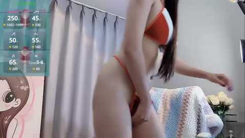 Media: Video of a slender, light-skinned woman in a red bikini, adjusting her thong in a bright, modern room with light gray curtains and a knitted throw on a sofa.