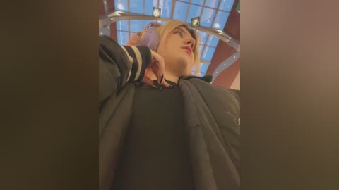 Media: Video of a blonde woman with a slim figure wearing a black jacket, taken from a low angle, showing a clear sky and glass ceiling.