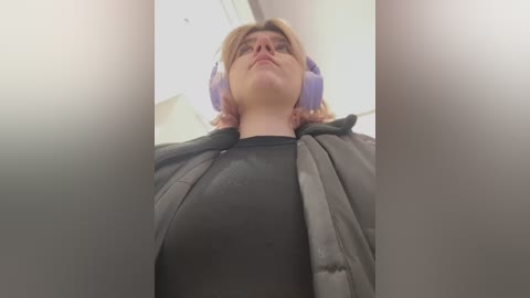 Media: Video of a woman with shoulder-length blonde hair wearing glasses, purple headphones, and a grey jacket over a black shirt, taken from a low angle, creating a dramatic effect.