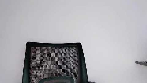 Media: Video of a black mesh office chair with a green cushion, positioned against a plain white wall. The chair is placed centrally in the frame, slightly tilted, with a minimalist setting.