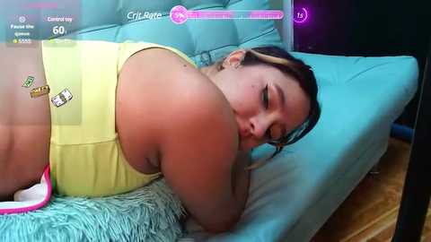 Media: Video of a plus-size woman with light brown skin, wearing yellow shorts and a tank top, lying on a blue couch with a pink pillow.