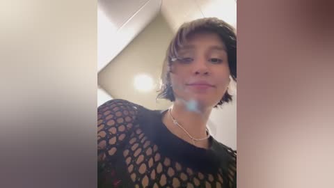 Media: Video of a woman with short, wavy brown hair, wearing a black, fishnet top and pearl necklace, indoors with blurred, white ceiling and walls.