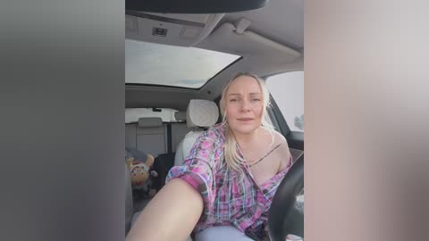 Media: Video of a smiling blonde woman in a colorful plaid shirt, seated in a car with beige interior, with a plush toy visible in the background.