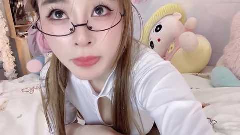 Media: A video of an Asian woman with long brown hair and glasses, wearing a white shirt, kneeling on a bed. Background includes plush toys, pastel-colored bedding, and a soft, dreamy atmosphere.