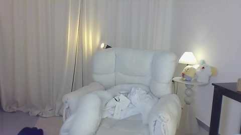 Media: Video of a white leather recliner chair with a white blanket draped over it, positioned against white curtains. A small white lamp with a shade is on a side table beside the chair.