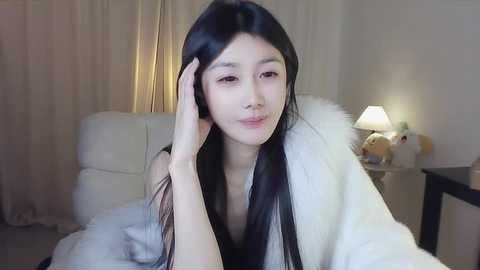 Media: Video of a young East Asian woman with long black hair, fair skin, and a slender build, resting her hand on her cheek. She's wearing a white bathrobe in a softly lit bedroom with a white bed, beige curtains, and a nightstand with a lamp and stuffed toys.