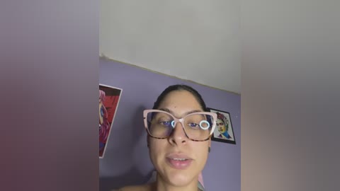 Video of a young woman with light skin, straight dark hair, wearing large, pink-framed glasses, standing in a room with a purple wall, a poster, and a framed picture.