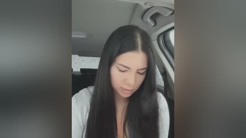 Media: Video of a young woman with long, straight black hair, fair skin, and a slender physique, wearing a white top. She appears contemplative inside a car, with a gray interior and a partially visible driver's seat.