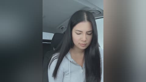 Media: Video of a young woman with long black hair, fair skin, and light makeup, wearing a light blue shirt, sitting in a car, looking downward.