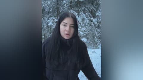 Media: Video of a young Asian woman with long black hair, wearing a black fur coat, standing in a snowy forest with snow-covered trees in the background.