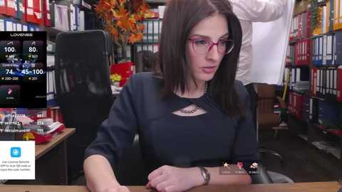 Media: Video of a woman with straight brown hair, red-rimmed glasses, and a black blouse, sitting at a desk in a library-like setting.