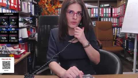 Media: Video of a brunette woman with glasses and a black blazer, seated at a cluttered desk in a library, holding a phone, surrounded by colorful bookshelves.