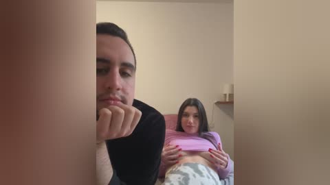 Media: A video of a young man and woman in a bedroom. The man, with short dark hair, is leaning against a pillar, looking pensive. The woman, with long dark hair, is lying in bed, wearing a pink shirt and gray pajama pants.