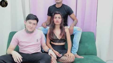 Media: Video of three people on a green couch: a man in a pink shirt and black pants, a tattooed woman in a black lace bra and fishnet stockings, and a shirtless man in jeans.