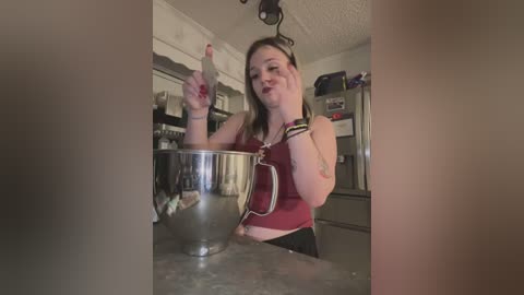 Media: Video of a young woman with medium build, wearing a red tank top, standing in a cluttered kitchen, pointing with her right hand while stirring a large pot on the stove.