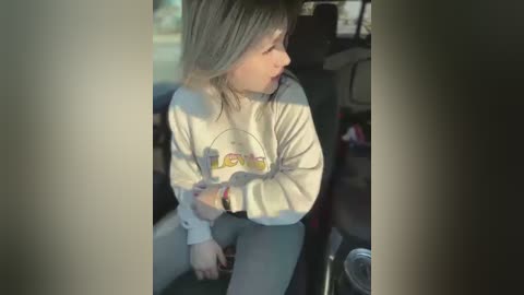 Media: Video of a young girl with light skin, shoulder-length blonde hair, wearing a cream-colored graphic sweatshirt and light pants, sitting in the backseat of a car. Sunlight streams in, casting shadows on her face.