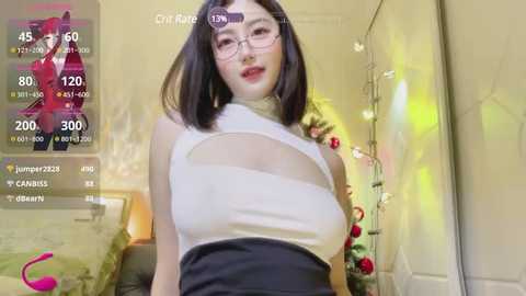 Media: Video of a slender, fair-skinned woman with shoulder-length black hair, wearing glasses and a white, high-cut top revealing her cleavage, standing in a softly lit bedroom with a Christmas tree in the background.