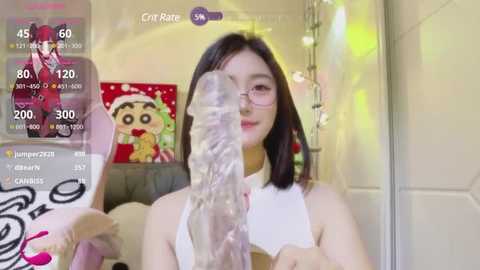 Media: Video of an East Asian woman with straight black hair and glasses, wearing a white top, holding a clear dildo. Background includes a Christmas-themed wall and a monitor displaying a live stream.