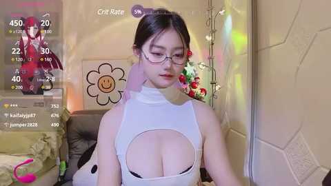 Media: Video of an Asian woman with dark hair, glasses, and a white, cut-out top revealing her ample breasts, sitting in a room with a colorful, cartoon-themed background.