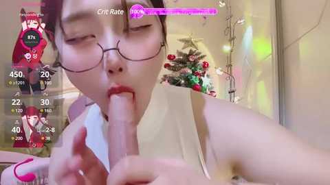 Media: A video of an Asian woman with glasses performing oral sex on a penis, wearing a white tank top, in a festive, decorated room with a Christmas tree.