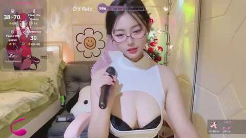 Media: A video of an East Asian woman with glasses, large breasts, wearing a revealing white top, holding a microphone, in a cozy, colorful bedroom.