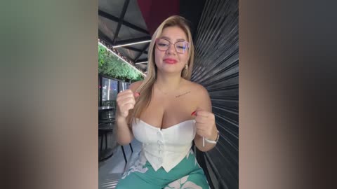 Media: Video of a blonde woman with glasses, fair skin, wearing a white corset and green floral dress, sitting indoors near a black shutter, with a green plant in the background.