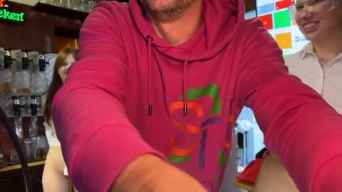 Media: A video shows a person wearing a bright pink hoodie with colorful geometric patterns, smiling and holding a drink. The background features a bar with bottles and a smiling woman in a white shirt.