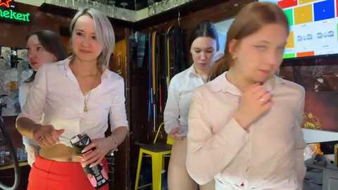 Media: Video of four women in a lively, cluttered room. Two women in white shirts and red skirts stand, holding bowls and smiling. Background shows colorful posters, wooden shelves, and a TV.