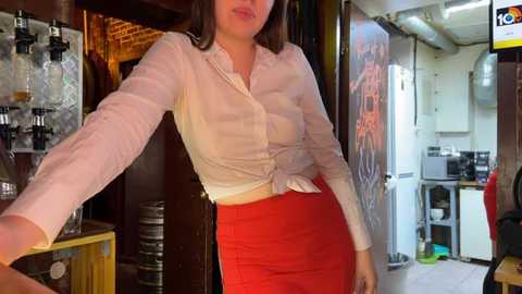 Media: Video of a woman in a white blouse, tied at the waist, and red skirt, leaning against a wooden bar in a rustic, dimly-lit pub.