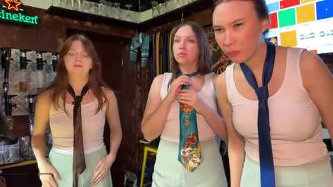 Media: Video of three young women in a bar, wearing sleeveless tops and ties, one with a medal, in a dimly lit, cluttered setting.