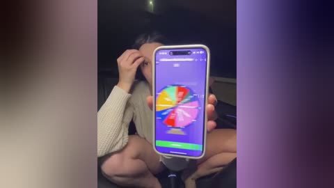 Media: Video of a person with dark hair, wearing a beige sweater, sitting cross-legged on a dark floor, taking a selfie with a smartphone displaying a colorful, abstract background.