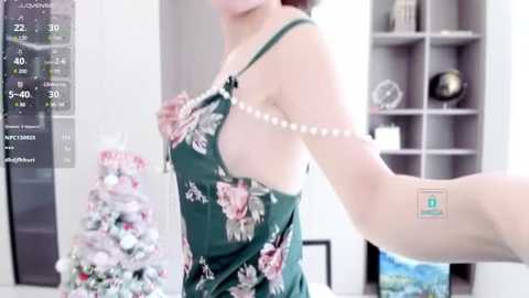 Media: A video of a fair-skinned woman in a green floral sleeveless top, holding a phone displaying a weather app, standing indoors beside a decorated Christmas tree and shelves.