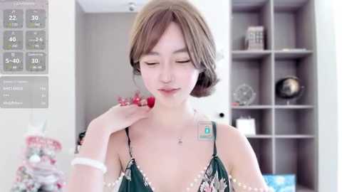 Media: A video of an Asian woman with light skin and short brown hair, wearing a green floral dress, eating a strawberry, standing in a modern, minimalist room with shelves and a calendar.