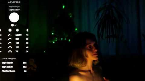 Media: A video of a woman with shoulder-length blonde hair, partially illuminated, standing in a dimly lit room with a Christmas tree and green lights in the background.