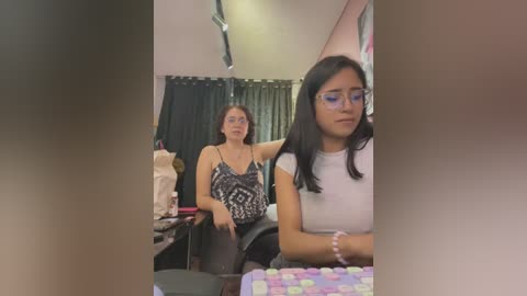 Media: Video: A woman with glasses and long black hair, wearing a white top, sits at a desk in a dimly lit room. Behind her, a sewing machine and sewing supplies are visible.