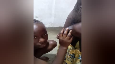 Media: A video shows a young African boy with dark skin, sitting with his hands on his knees, wearing a yellow shirt with a floral pattern. He appears in a dimly lit room with a white curtain and a beige wall.