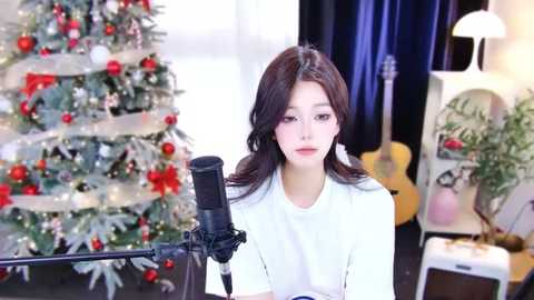 Media: Video of a young woman with long dark hair, fair skin, wearing a white t-shirt, singing into a microphone in front of a decorated Christmas tree with red ornaments and a guitar.