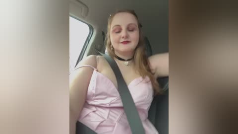 Media: Video of a smiling, fair-skinned woman with long blonde hair, wearing a pink strapless dress, seated in a car with a gray seatbelt, her arms relaxed.