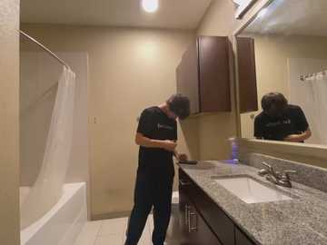 Media: Video of a man with short brown hair in a dark T-shirt and jeans, standing in a beige-tiled bathroom with a granite countertop, mirror, and dark wooden cabinets.