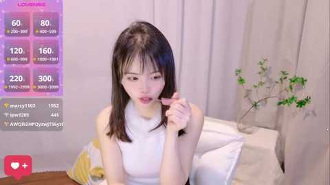 Media: A video of a young Asian woman with straight black hair and fair skin, wearing a sleeveless white top, eating a popsicle in a bedroom with white curtains and a green plant.