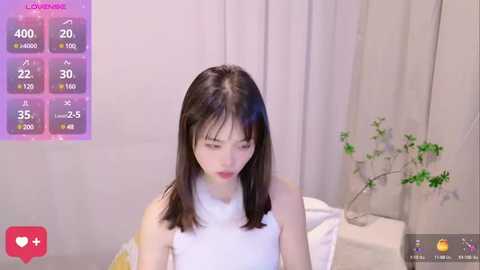 Media: A video of a young East Asian woman with straight black hair, wearing a white sleeveless top, sitting on a bed. Background features a plant, white curtains, and a digital screen displaying game statistics.