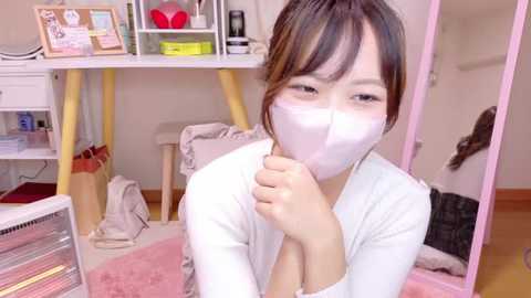 Media: Video of an Asian woman with a mask, sitting on a pink rug, in a cluttered bedroom with a white desk and a mirror.