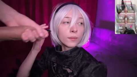 Media: Video of a pale-skinned, petite woman with short, platinum blonde hair and a black headband, wearing a dark, textured outfit, being combed by a hand. Background: deep red curtains, purple lighting, and a screen displaying a video feed.