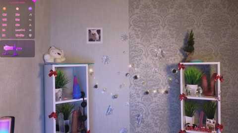 Video of a room with white wall, grey damask wallpaper, white shelves adorned with green plants, red bows, and festive decorations; a polar bear toy on the left.