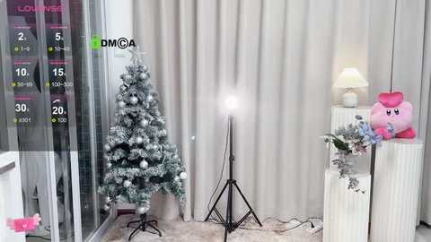 Media: A video of a minimalist, modern living room with a white Christmas tree, a pink plush Kirby toy, and a spotlight on a tripod, set against beige curtains and a white floor lamp.