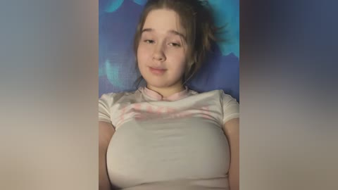 Video of a young woman with light skin, brown hair in a ponytail, wearing a tight, white t-shirt that accentuates her large breasts, against a blue and purple gradient background.