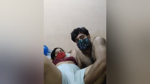 Media: Video of a man and woman in a small, dimly lit room. The man, shirtless, is holding the woman, who is gagged and bound. Both wear masks. The woman's face is red and she appears distressed.