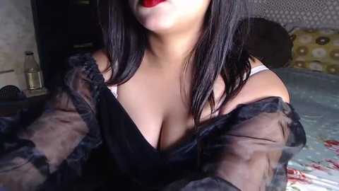 Media: A video of a woman with medium skin tone, straight black hair, and bold red lipstick, wearing a sheer black dress, revealing a white bra and cleavage. Background features a floral bedspread and dark room.